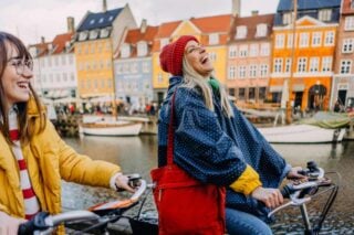 Two women laughing and cycling in Denmark, enjoying exercise and sightseeing, promoting a healthy lifestyle while abroad as part of their international healthcare goals