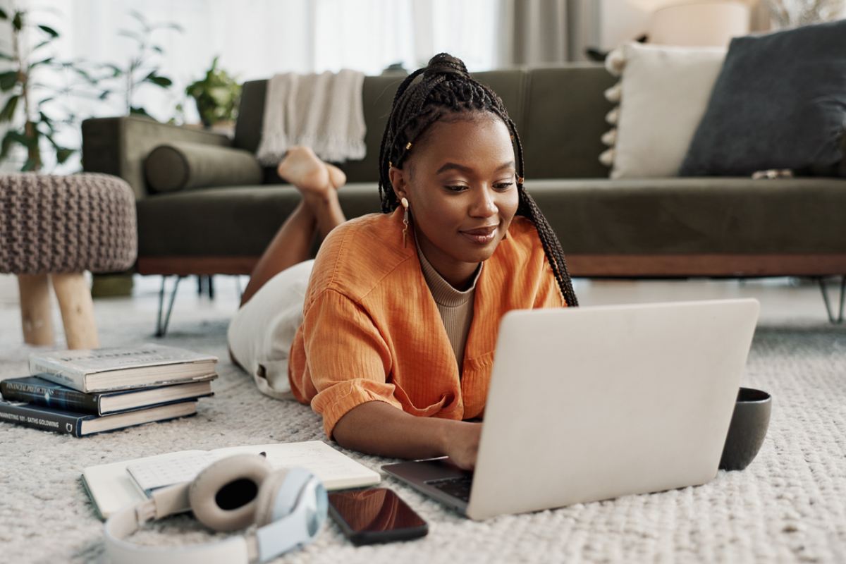 a young black woman on the floor of a living room looking down at her laptop as she researches options for studying abroad