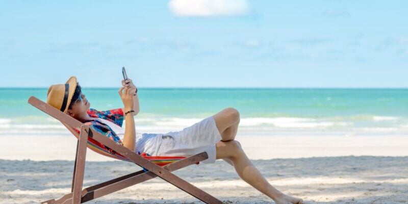 a male expat relaxing in a hammock and using his cell phone on a tropical beach in Thailand, researching the best international credit cards for living abroad