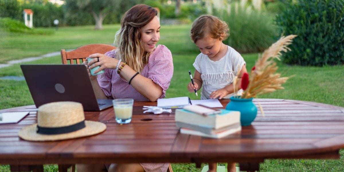 a mother working on her laptop in a garden as her child plays next to her