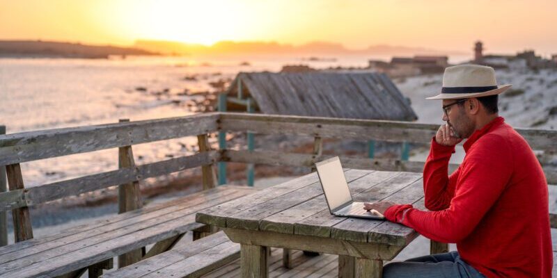 digital nomad sitting outdoors on the beach with a laptop alone doing telecommuting at sunset in brazil