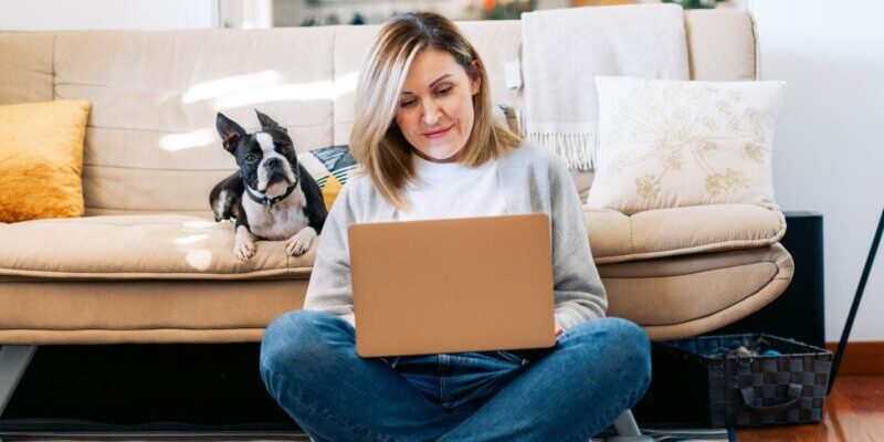 a female expat working remotely from home on her laptop with her dog sitting on the couch behind her