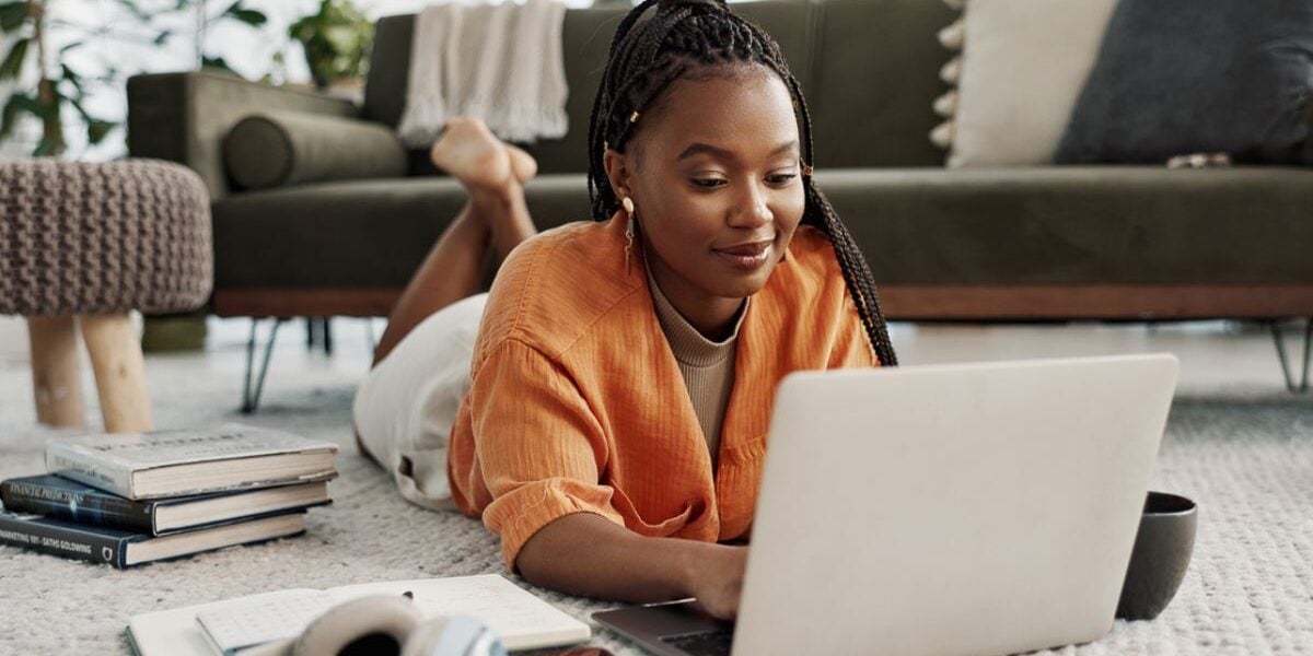 a young black woman on the floor of a living room looking down at her laptop as she researches options for studying abroad